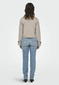 Veste beige courte avec des manches longues, des poignets boutonnés, et une fente au dos, portée avec un jean bleu clair et des bottes à talons noires.