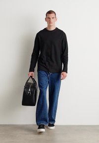 Camisa negra de manga larga, vaqueros azules holgados, bolso negro con asas y zapatillas negras con suelas blancas sobre un fondo liso.