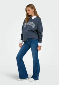 Felpa oversized grigia scura con la scritta "LOS ANGELES CALIFORNIA", abbinata a jeans a zampa d'elefante blu e scarpe da ginnastica bianche. Camicia bianca con colletto visibile.