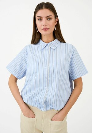 Žena v svetlomodrej pruhovanej košeli s krátkym rukávom a predným zipsom, béžových nohaviciach a kruhových náušniciach stojí s rukami v vreckách.