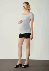 Grå ammebluse med korte ermer og samlede sider, kombinert med svarte shorts. Modellen har på seg svarte sandaler med tykke hæler. Nøytral bakgrunn.