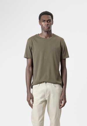 Hombre de pie mirando hacia adelante, vestido con una camiseta verde oliva y pantalones beige claros contra un fondo blanco simple.