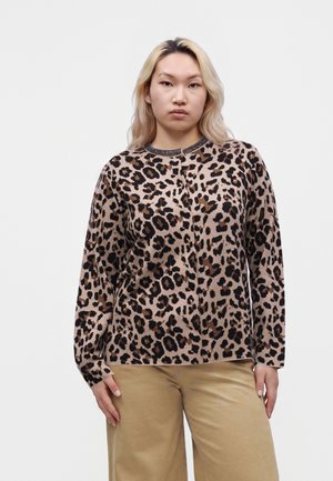 Jeune femme aux longs cheveux blonds portant un haut à manches longues imprimé léopard et un pantalon beige, debout sur un fond blanc.