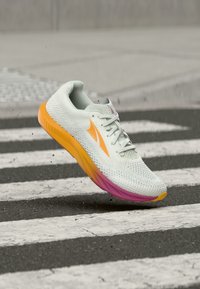 Λευκό αθλητικό παπούτσι με πορτοκαλί και ροζ σόλα και λογότυπο, αιωρούμενο πάνω από μια ασπρόμαυρη διάβαση πεζών σε δρόμο.