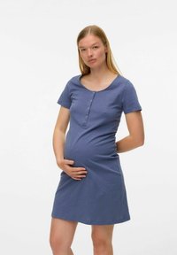 Mujer embarazada de pie con una mano en su barriga y la otra detrás de su espalda, vistiendo un vestido de botones azul de manga corta.