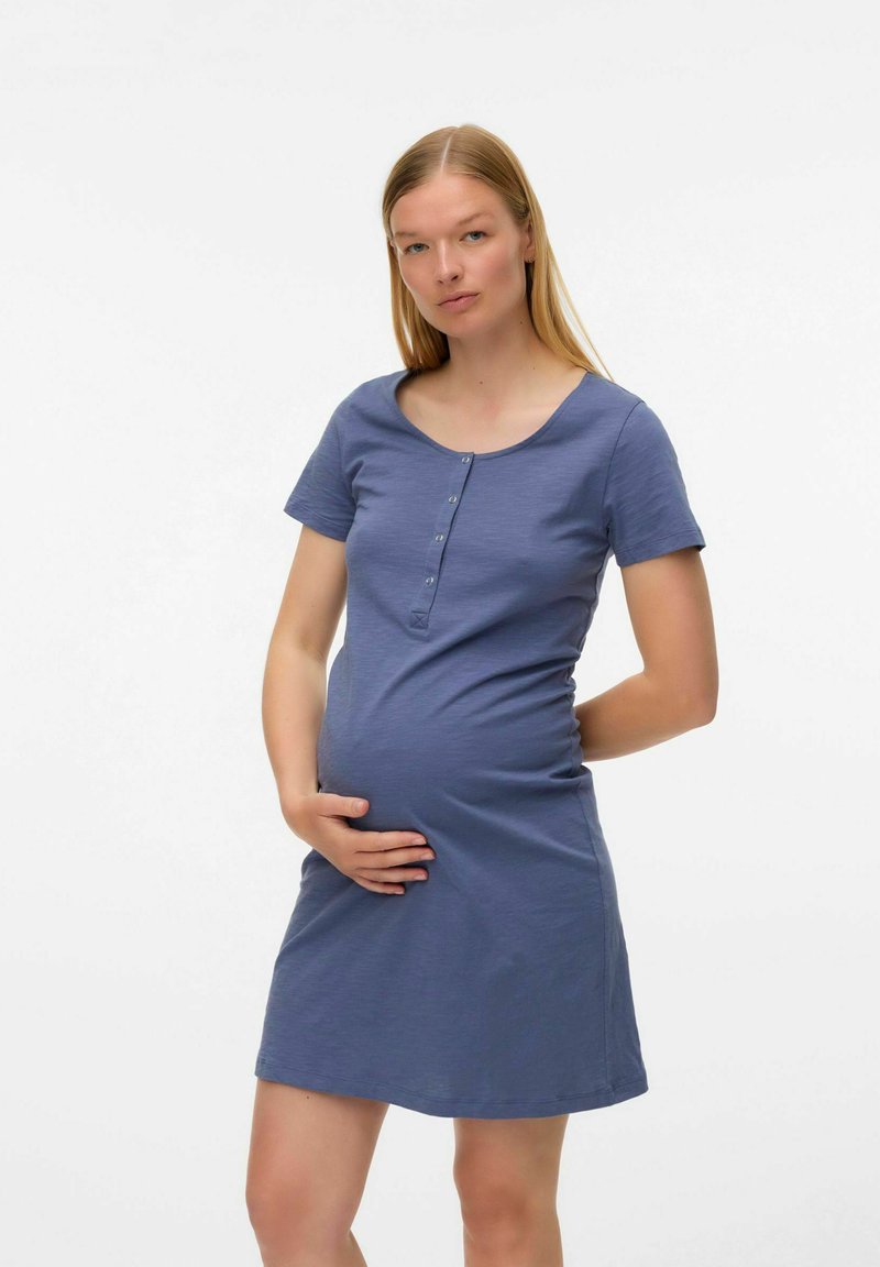 Mujer embarazada de pie con una mano en su barriga y la otra detrás de su espalda, vistiendo un vestido de botones azul de manga corta.