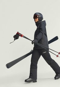 Lyžiarske vybavenie pozostávajúce z čiernych lyžiarskych nohavíc a bundy, čiernych lyžiarskych topánok, červených akcentov na lyžiach, čiernych rukavíc a čiernej prilby so štítom.