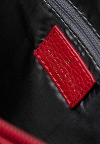 Etiqueta de cuero texturizado rojo con el texto en relieve dorado "LAZAROTTI" en un interior de tela negra, junto con un zipper gris.