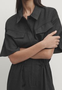 Dark gray utility-style shirt with short sleeves, two chest pockets, and a cinched waist. Material appears smooth with a subtle texture.