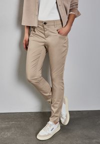 Pantalones beige ajustados de tejido suave, con bolsillos delanteros, cierre de botón y un ligero brillo, combinados con zapatillas blancas.