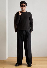 Černý pletený svetr, volně střižené černé kalhoty, hnědá bunda přehozená přes ruku a černé boty. Hladké textury a neutrální barevná paleta.