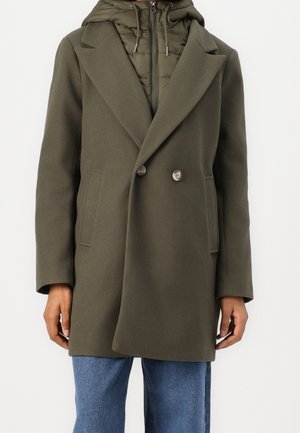 Personne portant un manteau croisé vert olive superposé à une veste matelassée zippée avec capuche de couleur vert olive et un jean en denim bleu.