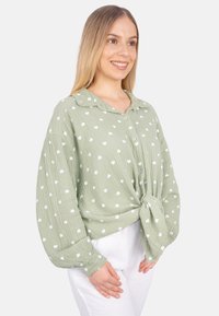 Green, button-up blouse with a collar, featuring white heart-shaped patterns, long sleeves, and a knotted front detail. Textured fabric.