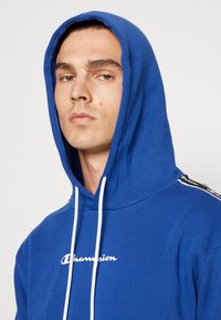 Felpa blu realizzata in morbido tessuto con un logo bianco sul petto. Presenta un cappuccio con coulisse e accenti laterali neri a contrasto.