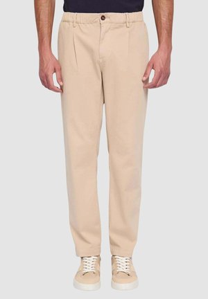 Pantaloni beige con vestibilità rilassata, caratterizzati da pieghe, cintura elasticizzata e chiusura con bottone. Abbinati a sneakers di colore chiaro.
