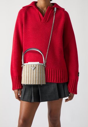 Červený pletený svetr s výstřihem do V, doplněný černou kostkovanou plisovanou sukní. Malá tkaná kabelka v krémové a zlaté barvě je držena na stříbrném řetízku.