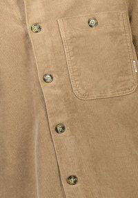 Camisa de pana marrón con estrías verticales, que cuenta con cuatro botones de latón y un bolsillo en el pecho. Textura suave y diseño resistente.