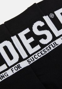 Calcetines negros con letras de marca blancas "DIESEL" en la parte superior. Presentan una textura acanalada y un acabado suave. Contienen una banda de texto secundaria debajo.
