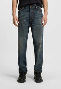 Donkerblauwe denim jeans met een ontspannen pasvorm, zichtbare naden en lichte vervaging op de dijen. Gecombineerd met zwarte sneakers voor contrast.