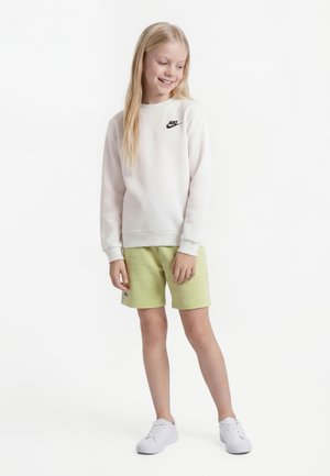 Pige med langt blondt hår iført en hvid Nike-sweatshirt, lysegrønne shorts og hvide sneakers, stående og smilende, kiggende mod venstre.
