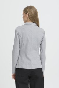 Vrouw met lang haar die een lichtgrijs aansluitend colbert en een zwarte broek draagt, van achteren gezien tegen een effen lichte achtergrond.