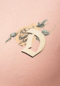 Έμπλαστρο με το γράμμα "D" σε εκρού χρώμα με μαύρο περίγραμμα πάνω σε εκτυπωμένο μπεζ τριαντάφυλλο με πράσινα φύλλα σε ροζ υφασμάτινο φόντο.