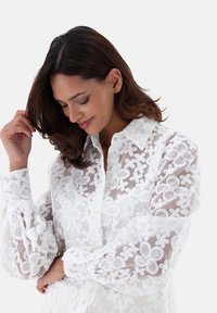 Sheer white shirt with floral embroidery, button-down front, and puffed sleeves, featuring intricate textured patterns and a collar.