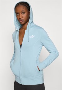 Ανοιχτό μπλε φούτερ με φερμουάρ, κατασκευασμένο από μαλακό ύφασμα, με κουκούλα, μπροστινές τσέπες και μικρό λευκό λογότυπο Puma στο στήθος.