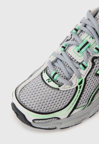Baskets de course en mesh gris avec des accents verts et noirs, présentant une surface texturée, un bout arrondi et une semelle en caoutchouc plate. Les lacets sont gris.