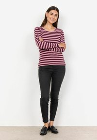 Maglietta a maniche lunghe a righe in rosso scuro e lavanda, abbinata a jeans neri slim-fit e mocassini neri, in piedi davanti a uno sfondo bianco.