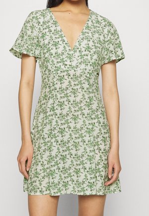 Žena v krátkým rukávem, šatech s výstřihem do V, s lehce krémovým podkladem a zeleným květinovým vzorem.