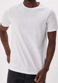 Mann trägt ein schlichtes weißes T-Shirt mit kurzen Ärmeln und schwarze Hose, steht mit einer Hand hinter dem Rücken und der anderen locker hängend.