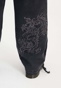 Pantalones de mezclilla negra con intrincados bordados de dragón gris en la parte inferior de la pierna. Incluyen un puño ajustado y un detalle de cordón cerca del tobillo.