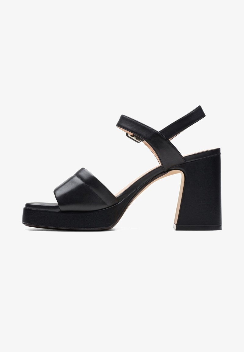 Sandale platform en cuir noir avec larges brides, boucle de cheville réglable et talon bloc haut et épais sur fond blanc.