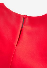 Gros plan du col d’un vêtement rouge avec un seul bouton rouge qui fixe une petite boucle en tissu.