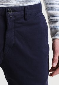 Pantalons en coton bleu marine avec une texture lisse, dotés d'une fermeture à bouton et de poches latérales, présentés avec un pull en tricot partiellement visible.