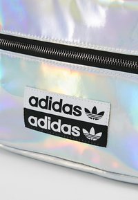 Mochila holográfica con una superficie brillante e iridiscente. Presenta un cierre negro y un destacado parche con el logo de "adidas" en negro y blanco.