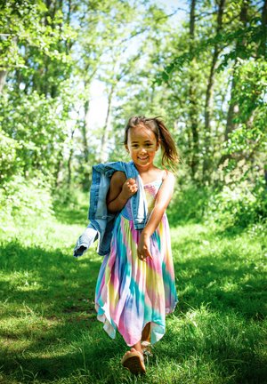 Pastellfarbendes Regenbogen-Maxikleid mit fließendem Design, Jeansjacke auf der Schulter, unterwegs auf einem grasbewachsenen Weg, umgeben von Bäumen.