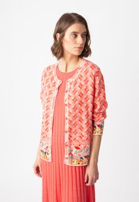 Röd mönstrad cardigan med knapplist, som har färgglada blommönster på ärmarna, bärs över en matchande texturerad rosa klänning.