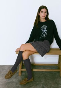 Suéter negro con un diseño de perro, falda gris con volantes, calcetas grises hasta la rodilla y zapatos de mocasín en color tostado. Fondo de banco de madera simple.