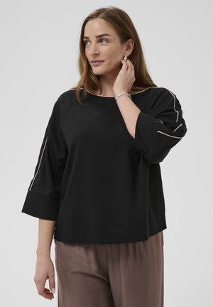 Femeie cu păr castaniu deschis purtând un top negru cu margini bej pe mâneci și pantaloni bej, atingând cerceii ei argintii.