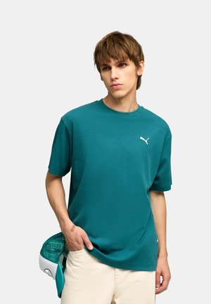 Młody mężczyzna w turkusowej koszulce Puma, trzymający pasującą turkusowo-białą czapkę, stojący na tle jednolitego, jasnego tła.