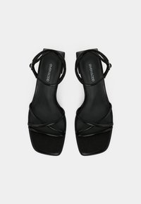 Black strappy sandals with a square toe, featuring thin crossed straps and a soft textured sole. Brand logo visible on the insole.