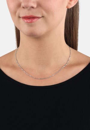 Vrouw in een dunne zilveren ketting boven een zwart geribd topje, met licht gekleurde lippen en haar achter de oren vastgebonden.