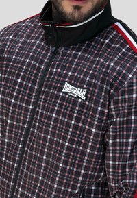 Czarna kurtka zapinana na zamek w kratę, z czerwonymi i białymi akcentami, logo Lonsdale na klatce piersiowej oraz wysoka stójka z teksturowanej tkaniny.