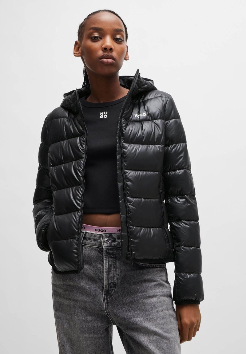 Schwarze Steppjacke mit Kapuze, glänzender Textur, gestepptem Design und Reißverschluss. Über einem schwarzen Oberteil mit Logo getragen; darunter sind graue Jeans sichtbar.