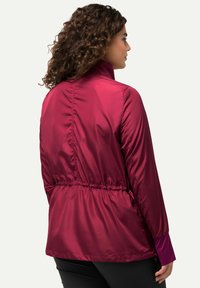 Lyhyt, viininpunainen takki, joka on valmistettu tasaisesta, kiiltävästä kankaasta. Tässä on vyötäröltä kireä malli, korkea kaulus ja joustavat hihansuut. Kevyt ja vartalonmyötäinen muotoilu.