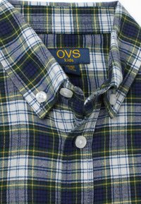 Camicia a quadri verde e blu navy con dettagli bianchi e gialli. Presenta un colletto e bottoni di plastica. L'etichetta mostra "OVS kids."