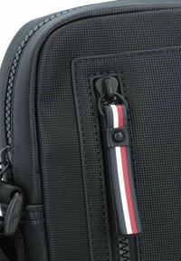 Zaino nero con texture, dotato di una cerniera liscia e un tirante con strisce rosse, bianche e blu. Materiale resistente con motivi sottili.