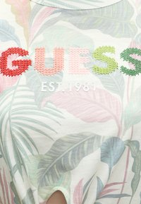 Έγχρωμη κεντημένη επιγραφή "GUESS" πάνω από το "EST. 1981" σε λευκό ύφασμα με παστέλ τροπικά φυλλαράκια.